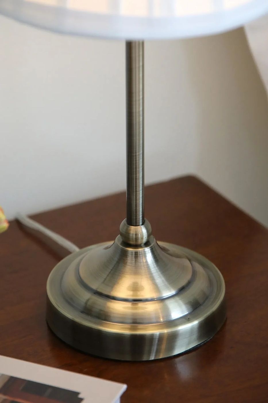 Lampa stołowa Oxford E14 w antycznym mosiądzu, z okrągłą, błyszczącą podstawą i prostym, metalowym trzonkiem. Ujęcie z boku, ukazujące detale podstawy i teksturę metalu, na tle drewnianego blatu. Kabel zasilający widoczny po lewej stronie.