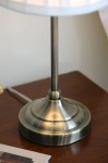 Lampa stołowa Oxford E14 w antycznym mosiądzu, z okrągłą, błyszczącą podstawą i prostym, metalowym trzonkiem. Ujęcie z boku, ukazujące detale podstawy i teksturę metalu, na tle drewnianego blatu. Kabel zasilający widoczny po lewej stronie.