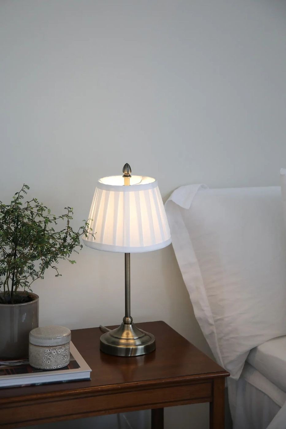 Lampa stołowa Oxford E14 w antycznym mosiądzu z białym, plisowanym abażurem. Ustawiona na drewnianym stoliku obok łóżka, z widoczną rośliną doniczkową i dekoracyjnym pojemnikiem. Kąt ujęcia frontalny, ukazujący detale lampy i otoczenie.
