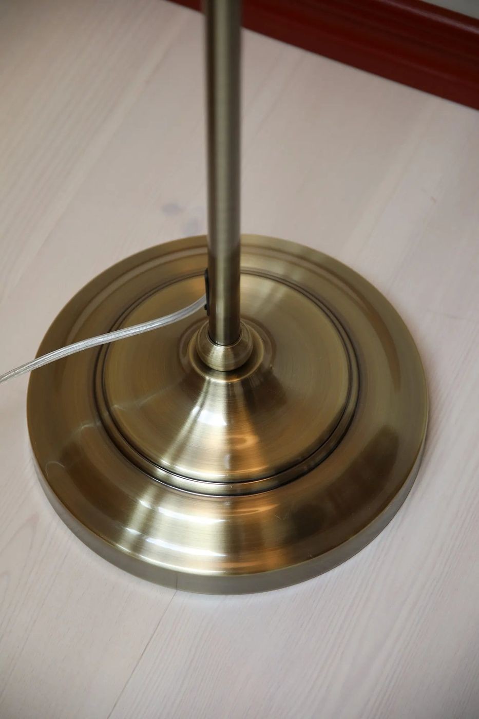 Lampa stołowa Oxford E14 w kolorze antycznego mosiądzu, z okrągłą podstawą. Widoczny metalowy słup z detalami wykończenia. Przewód elektryczny w kolorze przezroczystym, biegnący obok podstawy. Ujęcie z dołu, prezentujące szczegóły podstawy.