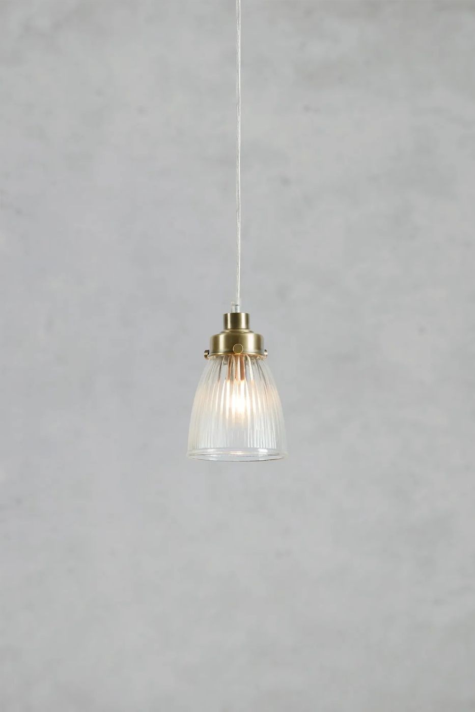 Lampa wisząca Bellis E14 w antycznym mosiądzu, z szklanym kloszem w pionowe żłobienia. Zawieszona na cienkim przewodzie, emitująca ciepłe światło. Klosz o zaokrąglonym kształcie, z widocznym źródłem światła wewnątrz. Tło jasnoszare.