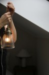 Lampa wisząca Bellis E14 w kolorze antycznego mosiądzu z przezroczystym, żłobkowanym kloszem. Ujęcie z bliska, ręka trzymająca lampę, która emituje ciepłe światło. Tło neutralne, z widoczną lampą stołową w pomieszczeniu.