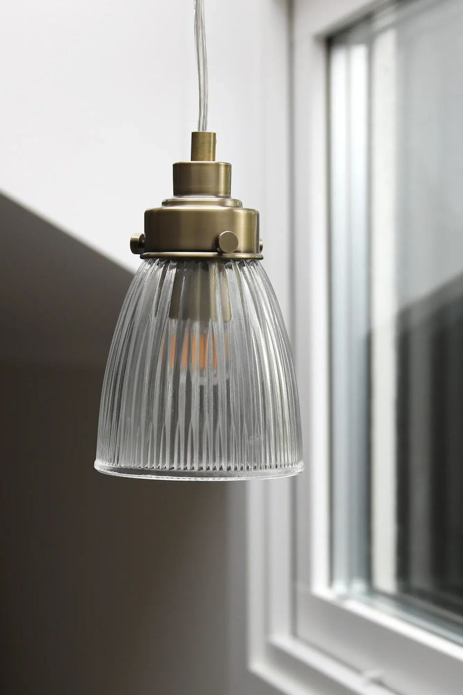 Lampa wisząca Bellis E14 w antycznym mosiądzu z przezroczystym, fakturowanym kloszem. Ujęcie z boku, widoczny detal oprawy oraz żarówki. Klosz ma kształt stożka z wyraźnymi żłobieniami, zawieszony na przezroczystym przewodzie.