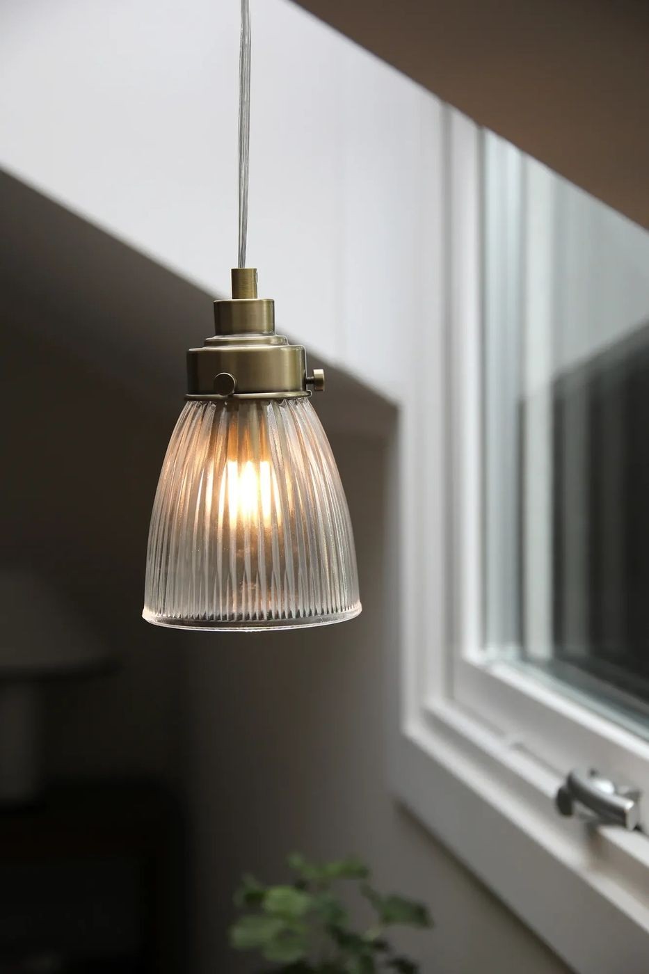 Lampa wisząca Bellis E14 w antycznym mosiądzu, z przezroczystym kloszem o żłobionej fakturze. Ujęcie z boku, wisząca na cienkim przewodzie. Widoczna ciepła żarówka wewnątrz klosza, otoczenie jasne, z oknem w tle.