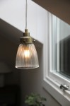 Lampa wisząca Bellis E14 w antycznym mosiądzu, z przezroczystym kloszem o żłobionej fakturze. Ujęcie z boku, wisząca na cienkim przewodzie. Widoczna ciepła żarówka wewnątrz klosza, otoczenie jasne, z oknem w tle.