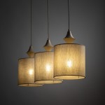 Lampa wisząca Amura w beżowym kolorze z trzema kloszami z lnu, zawieszona na długich przewodach. Klosze mają cylindryczny kształt i emitują ciepłe światło, z ozdobnymi elementami na górze każdego z nich. Tło ciemnoszare.