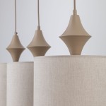 Beżowa lampa wisząca Amura z trzema kloszami z lnu, z widocznymi mocowaniami w kształcie stożka. Klosze mają teksturowaną powierzchnię, a całość zawieszona jest na cienkich przewodach. Ujęcie z przodu, pokazujące szczegóły konstrukcji.