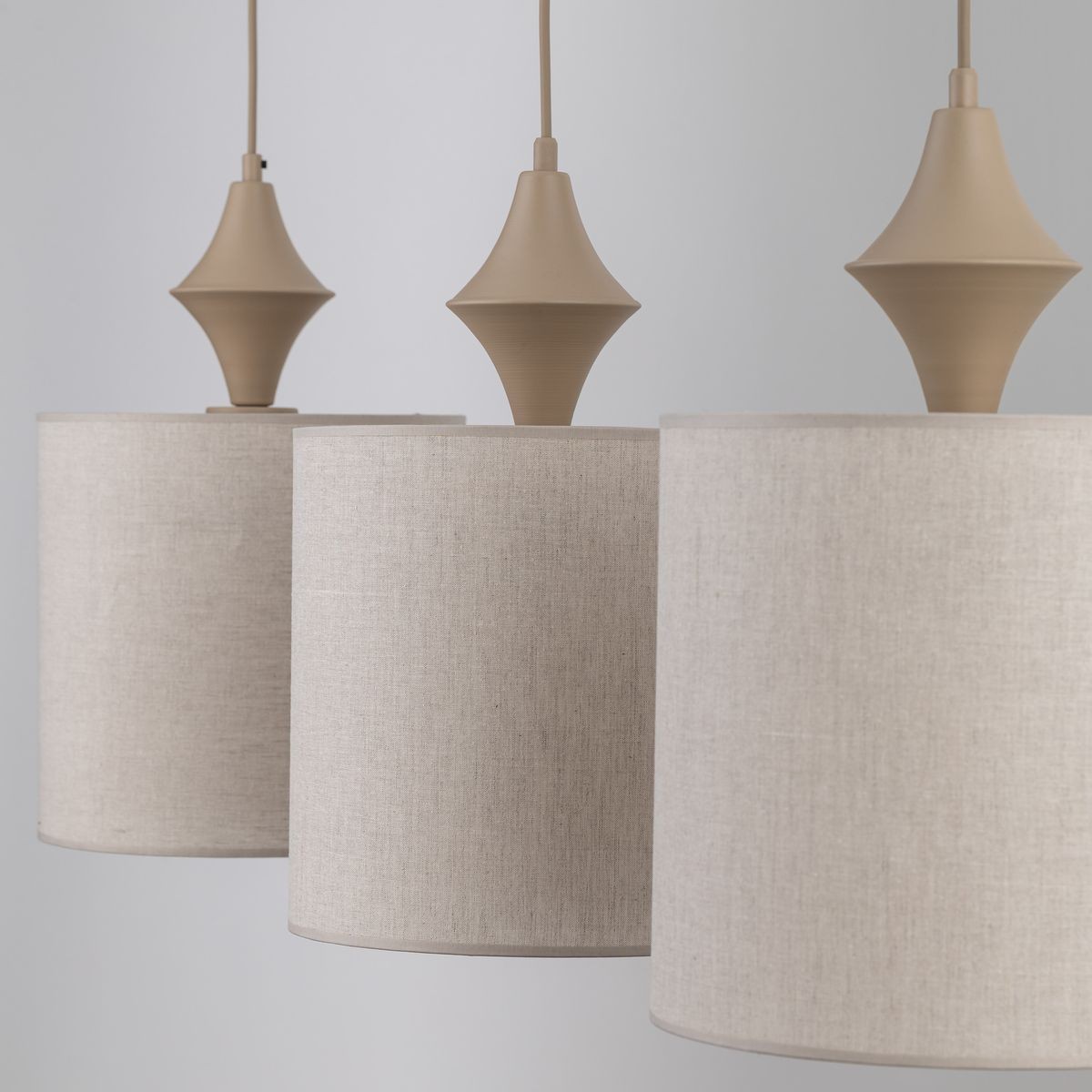 Lampa wisząca Amura w kolorze beżowym, składająca się z trzech cylindrycznych kloszy z lnu oraz ozdobnych elementów w kształcie diamentu. Klosze zawieszone na cienkich kablach, o neutralnym tle. Wymiary i detale wykończenia mogą się różnić.