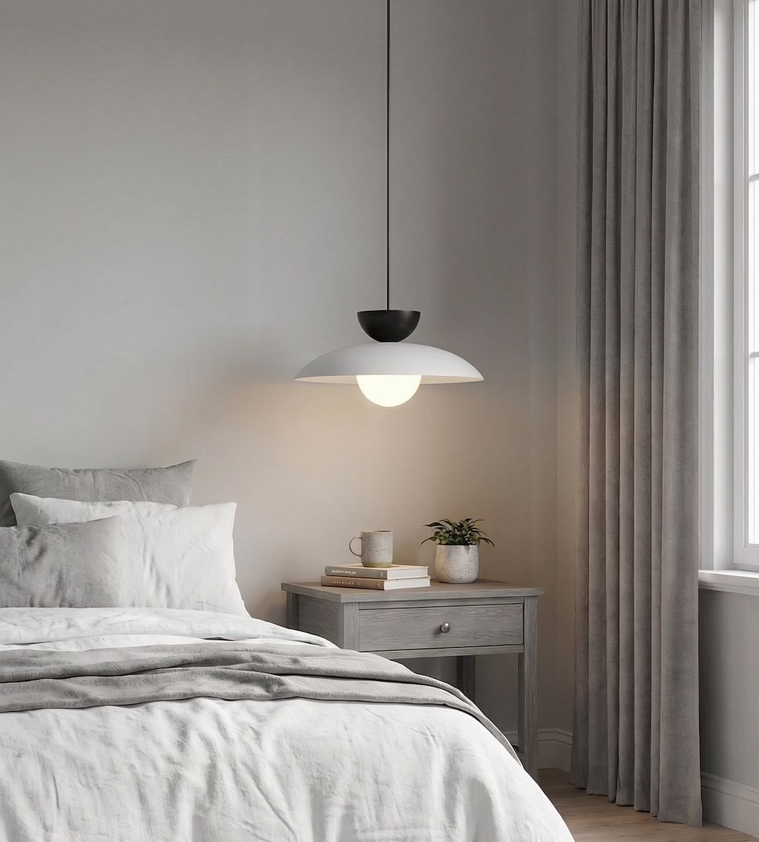 Lampa wisząca Mulan 1xG9 w kolorze szarym i czarnym, o zaokrąglonym kloszu w białym kolorze, zawieszona nad łóżkiem w sypialni. Widoczne elementy to szare zasłony, drewniana szafka nocna oraz roślina w doniczce.
