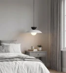 Lampa wisząca Mulan 1xG9 w kolorze szarym i czarnym, o zaokrąglonym kloszu w białym kolorze, zawieszona nad łóżkiem w sypialni. Widoczne elementy to szare zasłony, drewniana szafka nocna oraz roślina w doniczce.