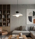 Lampa wisząca Mulan w kolorze szarym i czarnym z jednym źródłem światła G9, umieszczona nad stolikiem w nowoczesnym wnętrzu. Okrągła, matowa czasza z białym wykończeniem, zawieszona na cienkim, czarnym przewodzie. W tle drewniana półka z książkami.