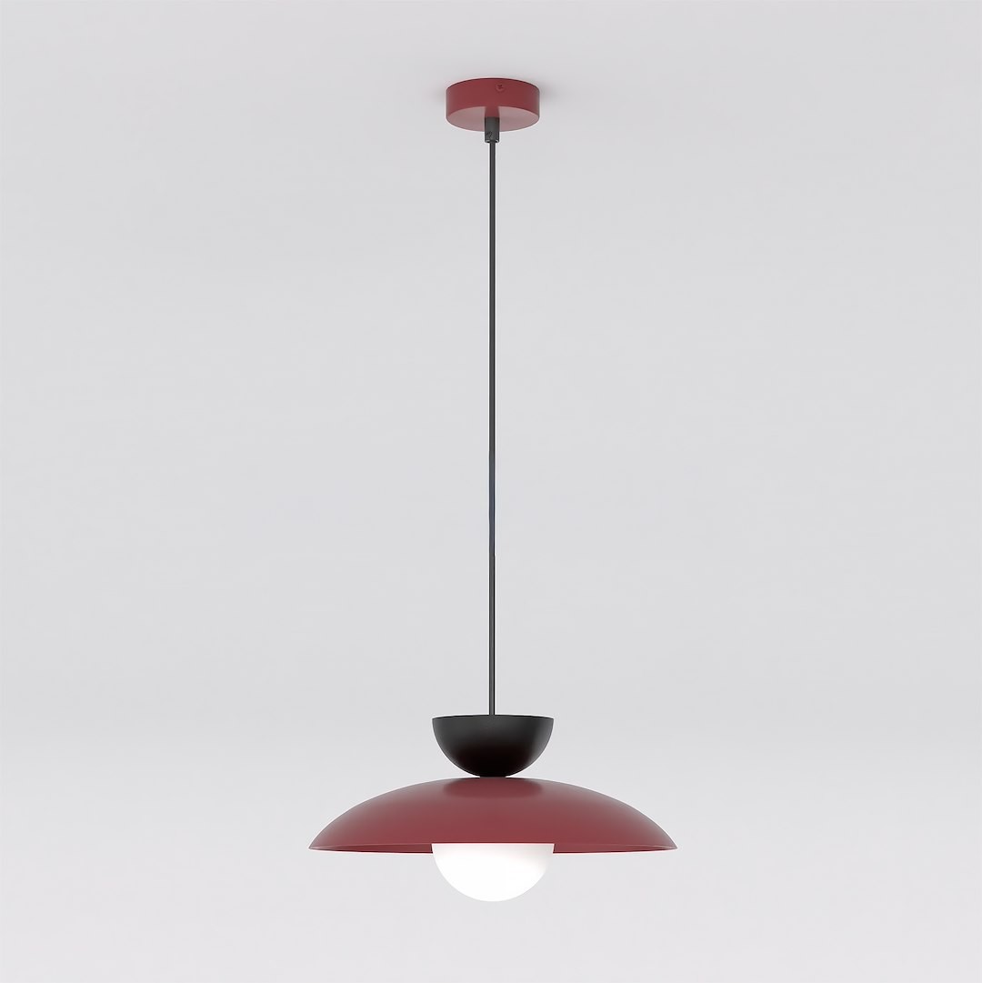Lampa wisząca Mulan w kolorze burgundowym z czarnym elementem, z jedną oprawą na żarówkę G9. Zawieszona na cienkim, czarnym przewodzie, z kulistym szklanym kloszem, umieszczona pod półkolistym metalowym daszkiem. Prosta forma na jasnym tle.