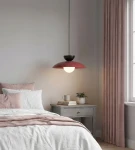 Lampa wisząca Mulan w kolorze burgundowym i czarnym, z okrągłym kloszem z białego szkła, zawieszona nad łóżkiem w sypialni. Obok stolik nocny z książkami i rośliną, na tle jasnej ściany i zasłon w odcieniu różowym.