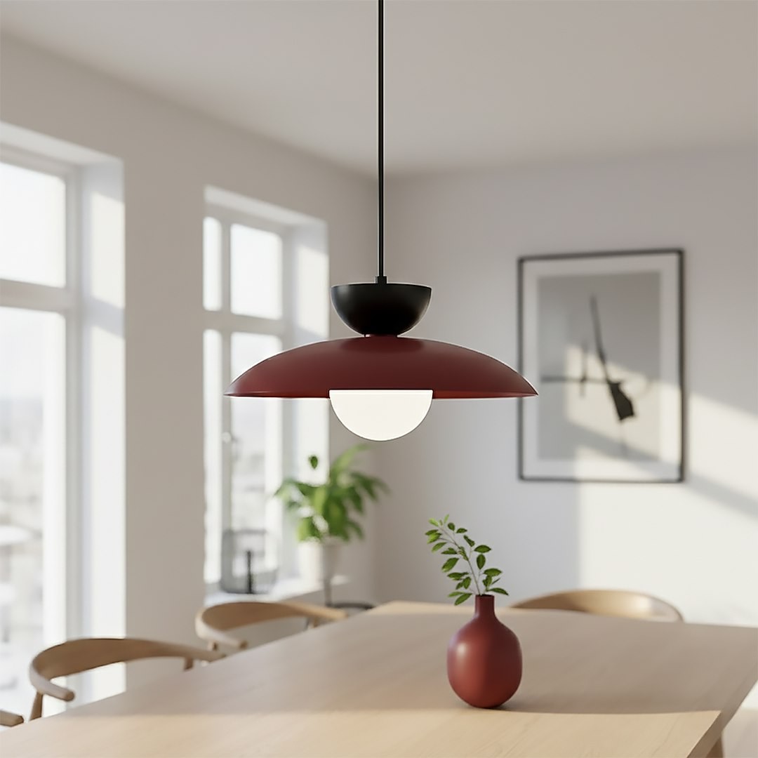 Lampa wisząca Mulan 1xG9 w kolorze burgundowym z czarnym uchwytem. Posiada okrągłą, białą oprawę świetlną. Zawieszona w nowoczesnym wnętrzu, na stole z drewnianym blatem, obok wazonu z rośliną.