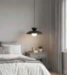 Lampa wisząca Mulan w kolorze czarnym z jedną oprawą G9, zawieszona nad łóżkiem w jasnej sypialni. Posiada okrągły klosz z przezroczystą częścią, umieszczona na tle szarych zasłon i drewnianej komody z rośliną.