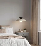 Lampa wisząca Mulan w kolorze mokka i czarnym, z pojedynczym źródłem światła G9. Zawieszona nad łóżkiem, z okrągłym kloszem w białym odcieniu i czarną osłoną. W tle beżowe zasłony i stół nocny z rośliną oraz filiżanką.