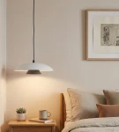 Lampa wisząca Babel 1xGX53 w kolorze czarnym i białym, o formie talerza, zawieszona na czarnym przewodzie. W tle widoczne przytulne wnętrze sypialni z drewnianą szafką nocną, filiżanką i rośliną.