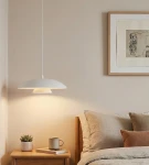 Biała lampa wisząca o okrągłym kształcie, zawieszona nad stolikiem nocnym. Na stoliku znajduje się filiżanka, mała roślina doniczkowa i książka. W tle widoczna ramka z grafiką. Ściana w neutralnym kolorze.