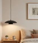 Lampa wisząca Babel 1xGX53 w kolorze czarnym z okrągłym kloszem, zawieszona nad stolikiem nocnym. W tle widoczne miękkie poduszki na łóżku oraz obraz na ścianie. Dodatki to filiżanka i roślina w doniczce.