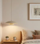 Lampa wisząca Babel 1xGX53 w kolorze mokka, o zaokrąglonym, płaskim kloszu, zawieszona nad stolikiem nocnym. Tło jasnoszare, z książkami, filiżanką i doniczką z rośliną. Stworzona do nowoczesnych wnętrz.