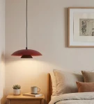 Lampa wisząca Babel 1xGX53 w kolorze burgundowym z czarnym kablem. Zawieszona nad stolikiem nocnym, o zaokrąglonym kształcie. W tle widoczna jasna ściana oraz elementy wystroju sypialni, takie jak poduszki i roślinność.