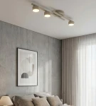 Fragment lampy sufitowej 3xGX53 w kolorze mokka, zamontowanej na suficie. Składa się z trzech okrągłych źródeł światła, skierowanych w dół, umieszczonych na poziomej listwie. W tle widoczna sypialnia z szarymi ścianami i jasnymi zasłonami.