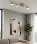 Fragment lampy sufitowej 3xGX53 w kolorze mokka, zamontowany na suficie, z trzema okrągłymi źródłami światła. Tło stanowi nowoczesny salon z obrazem i rośliną w doniczce.