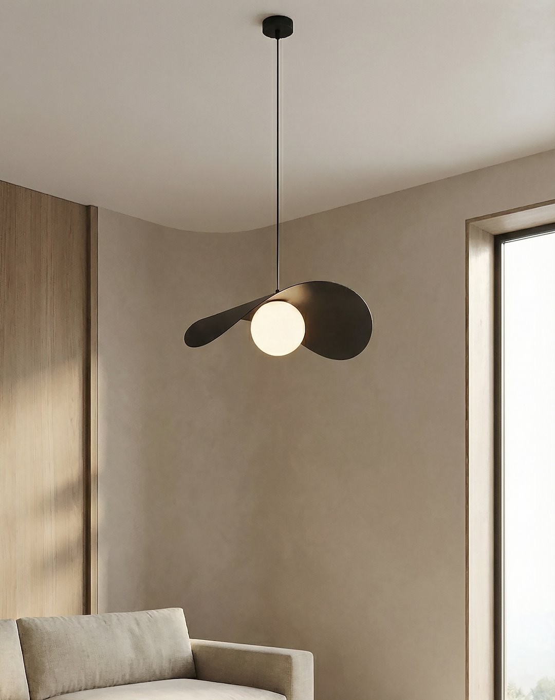 Lampa wisząca Ritual L z czarnym metalowym kloszem w kształcie liścia oraz okrągłą, białą żarówką G9. Zawieszona na cienkim, metalowym przewodzie, w nowoczesnym wnętrzu z sofą i dużym oknem.