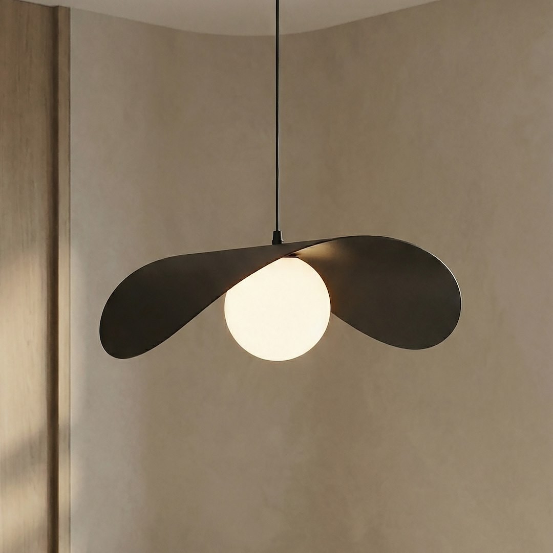 Czarna lampa wisząca Ritual L z jedną oprawą G9, z abażurem w kształcie dwóch skrzydełek, zawieszona na cienkim kablu. W centralnej części znajduje się okrągła, matowa żarówka, na tle jasnej, gładkiej ściany.