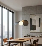 Czarna lampa wisząca Ritual L z jedną żarówką G9, zawieszona nad drewnianym stołem w nowoczesnym wnętrzu. Lampa ma minimalistyczny design z okrągłym kloszem i asymetrycznymi, płaskimi detalami. Tło stanowi betonowa ściana i duże okna.