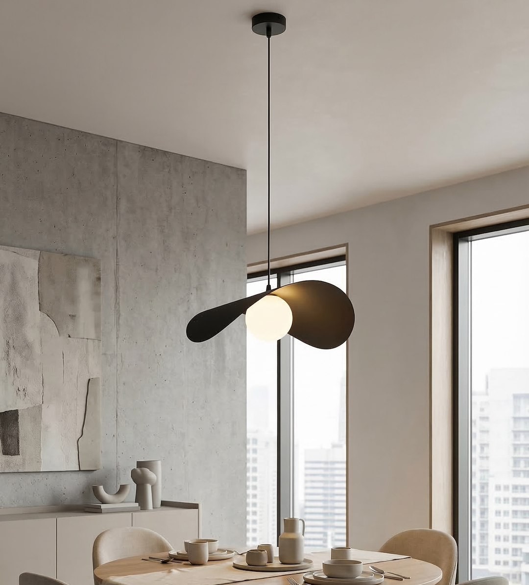 Czarna lampa wisząca Ritual L z jedną oprawą G9, o nowoczesnym kształcie z dwoma skrzydłami, umieszczona nad stołem w jasnym wnętrzu, z widokiem na okna. W tle betonowa ściana i drewniane meble.