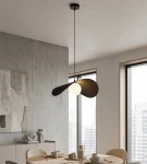 Czarna lampa wisząca Ritual L z jedną oprawą G9, o nowoczesnym kształcie z dwoma skrzydłami, umieszczona nad stołem w jasnym wnętrzu, z widokiem na okna. W tle betonowa ściana i drewniane meble.