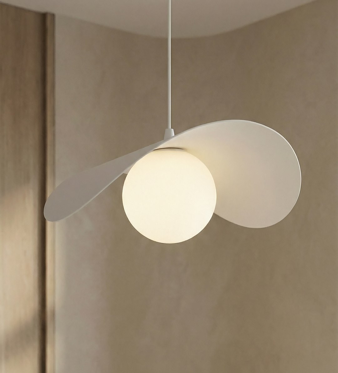 Lampa wisząca Ritual M w kolorze białym, z jedną oprawą na żarówkę G9. Okrągła, matowa kula świetlna otoczona jest dwoma asymetrycznymi, płaskimi elementami, zawieszona na cienkim przewodzie. Tło w neutralnym odcieniu.