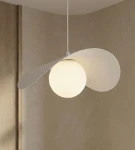 Lampa wisząca Ritual M w kolorze białym, z jedną oprawą na żarówkę G9. Okrągła, matowa kula świetlna otoczona jest dwoma asymetrycznymi, płaskimi elementami, zawieszona na cienkim przewodzie. Tło w neutralnym odcieniu.