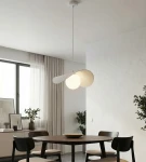 Lampa wisząca Ritual M w kolorze białym z jedną żarówką G9. Ma nowoczesny, opływowy kształt z dwoma falującymi elementami. Zawieszona na długim przewodzie w jasnym wnętrzu z drewnianym stołem i roślinnością w tle.