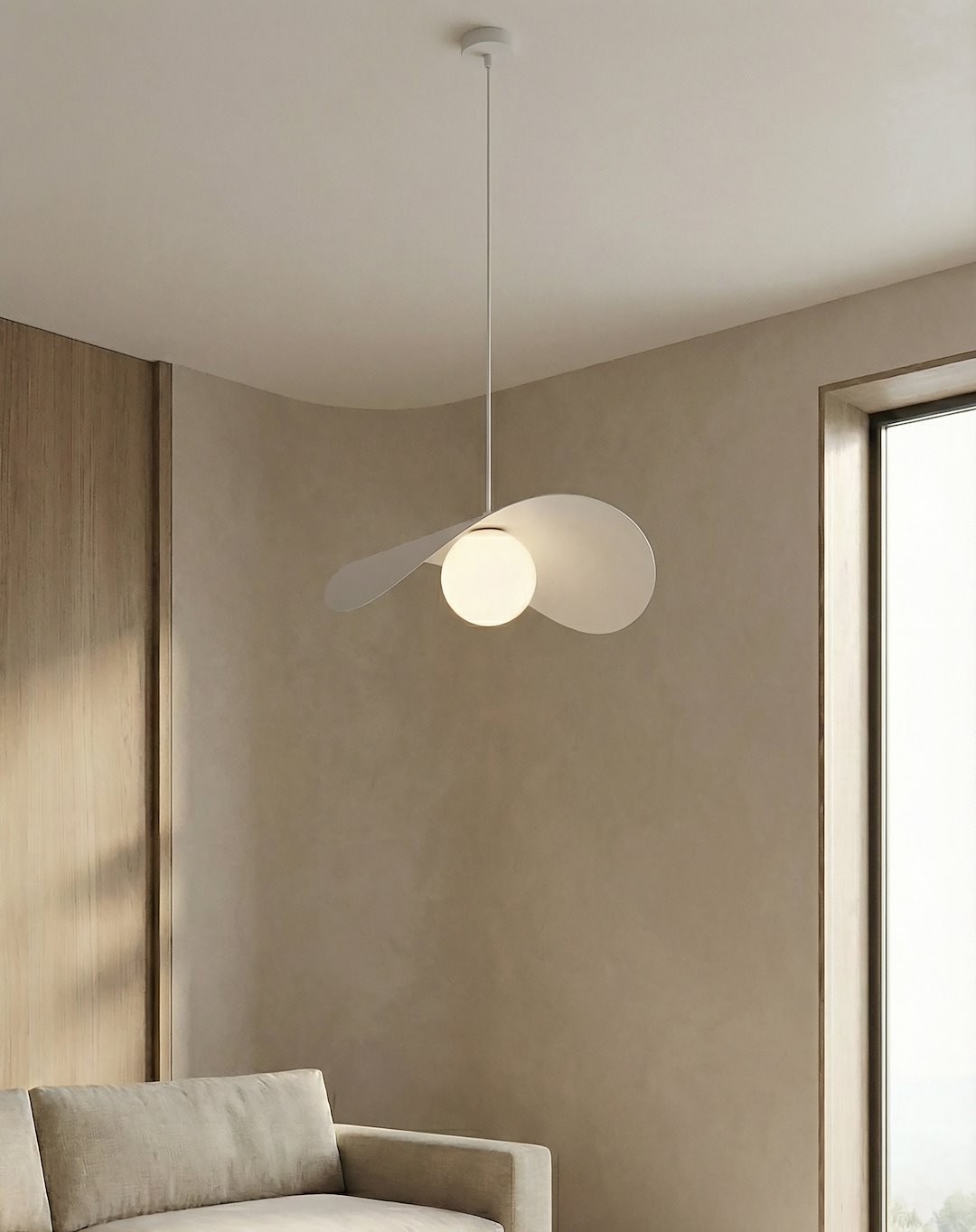 Lampa wisząca Ritual L w kolorze białym z pojedynczym źródłem światła G9, zawieszona na cienkim przewodzie. Posiada trzy asymetryczne, zaokrąglone elementy, tworzące nowoczesny design. Produkt w aranżacji wnętrza z jasnymi ścianami i kanapą.