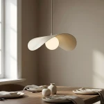 Lampa wisząca Ritual L w kolorze mokka z jednym źródłem światła G9. Posiada opływowy, dwuczęściowy klosz w odcieniu beżowym, zawieszony na cienkim metalowym przewodzie, nad drewnianym stołem z zastawą i ceramiką w jasnych kolorach.