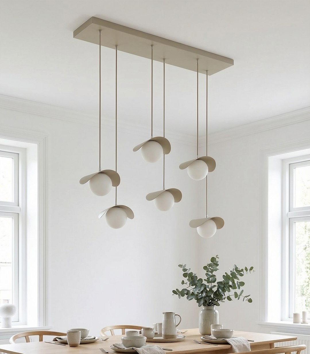 Lampa wisząca Ritual z sześcioma źródłami światła G9 w kolorze mokka, zawieszona na prostokątnej podstawie, z okrągłymi kloszami i dekoracyjnymi, opadającymi elementami. Umieszczona nad drewnianym stołem w jasnym wnętrzu.