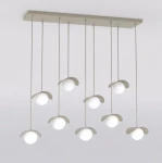 Lampa wisząca Ritual 9xG9 w kolorze mokka z dziewięcioma okrągłymi, białymi kloszami zawieszonymi na cienkich linkach. Ułożona równolegle do sufitu, z delikatnymi, zaokrąglonymi osłonami. Tło jasne, minimalistyczne.