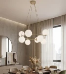 Lampa wisząca Azura 8xE14 w kolorze mokka z białymi, okrągłymi kloszami. Zawieszona na cienkich linkach, w centralnym punkcie pomieszczenia, w otoczeniu stołu z zastawą i dekoracjami. Tło z jasnymi zasłonami i betonowymi ścianami.