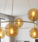 Lampa wisząca Azura w kolorze mokka z sześcioma bursztynowymi kloszami. Klosze w kształcie kul, zawieszone na okrągłej metalowej ramie. Oświetlenie z żarówkami E14, umieszczone w centralnej pozycji. Tło jasne, minimalistyczne wnętrze.