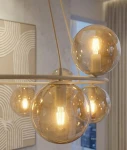 Lampa wisząca Azura z osiemnastoma żarówkami E14 w kolorze mokka i bursztynu. Posiada okrągłe, przezroczyste klosze, które emitują ciepłe światło. Zawieszona na cienkich kablach, w nowoczesnym wnętrzu, z jasnym tłem.
