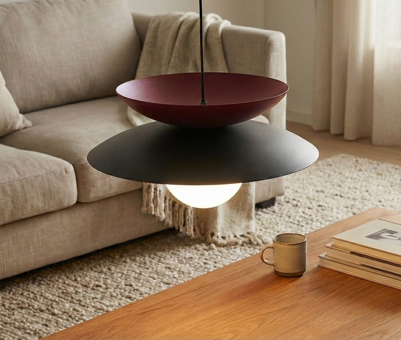 Czarna lampa wisząca Nagoya z jedną oprawą G9, posiadająca górną część w kolorze burgundowym i okrągłe, białe źródło światła. Zawieszona nad drewnianym stolikiem w jasnym wnętrzu, z widoczną sofą i dekoracyjną tkaniną w tle.