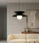 Czarna lampa wisząca Nagoya 1xG9 z okrągłym, białym kloszem, zawieszona na cienkim przewodzie. Widoczna w nowoczesnym wnętrzu z szarymi ścianami i jasną sofą. Kształt lampy podkreśla minimalistyczny styl.