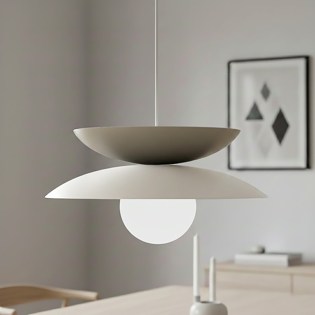 Lampa wisząca Nagoya z białym i mokka kloszem, o okrągłym, matowym kształcie, z centralną kulistą żarówką. Zawieszona na cienkim kablu w minimalistycznym wnętrzu, w tle widoczne drewniane meble i dekoracje.