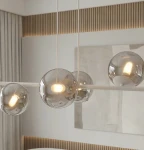 Lampa wisząca Magnus z 8 źródłami światła E14, w kolorze szarym i grafitowym. Posiada przezroczyste, kuliste klosze, zawieszone na białej konstrukcji. Widoczna w nowoczesnym wnętrzu z drewnianymi akcentami.
