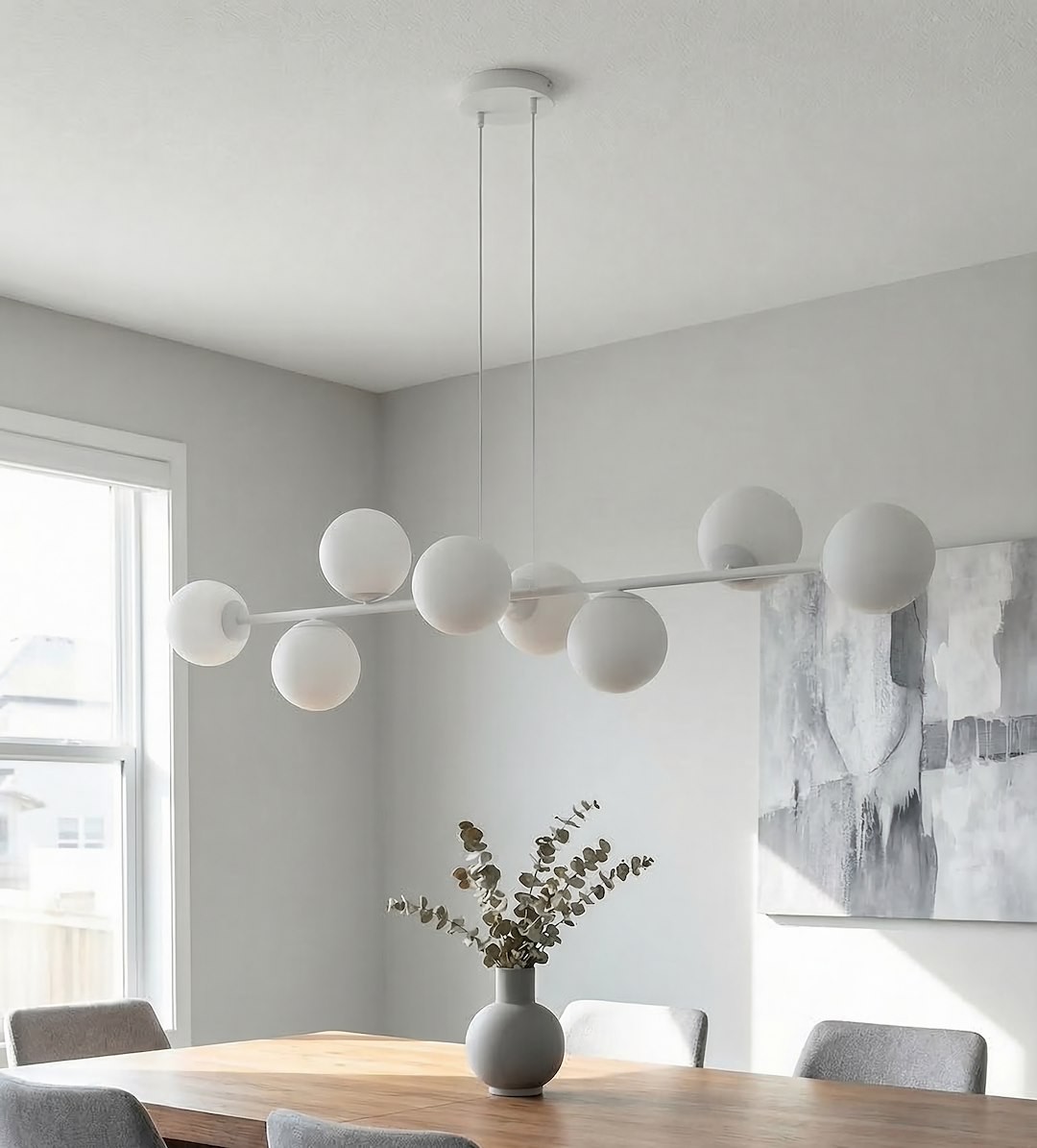 Lampa wisząca Magnus w kolorze szarym i białym z ośmioma kulistymi kloszami E14. Zawieszona na dwóch cienkich linkach, umieszczona nad drewnianym stołem. W tle widoczny obraz na ścianie oraz wazon z roślinami.