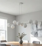 Lampa wisząca Magnus w kolorze szarym i białym z ośmioma kulistymi kloszami E14. Zawieszona na dwóch cienkich linkach, umieszczona nad drewnianym stołem. W tle widoczny obraz na ścianie oraz wazon z roślinami.