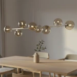 Lampa wisząca Magnus z 8 kulistymi kloszami w kolorze mokka i bursztynu, zawieszona nad drewnianym stołem. Klosze umieszczone na prostej, białej rurze, w tle widoczna wazon z gałązką. Jasne, minimalistyczne wnętrze.