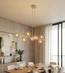Lampa wisząca Magnus 8xE14 w kolorze mokka i bursztynu, złożona z ośmiu szklanych kul, zawieszona nisko nad drewnianym stołem w nowoczesnej jadalni. Tło to betonowa ściana oraz zasłony, tworzące elegancką aranżację wnętrza.